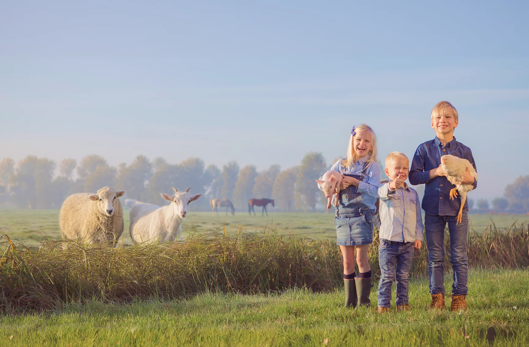 Drie kinderen poseren in de wei met op de achtergrond een schaap en geit
